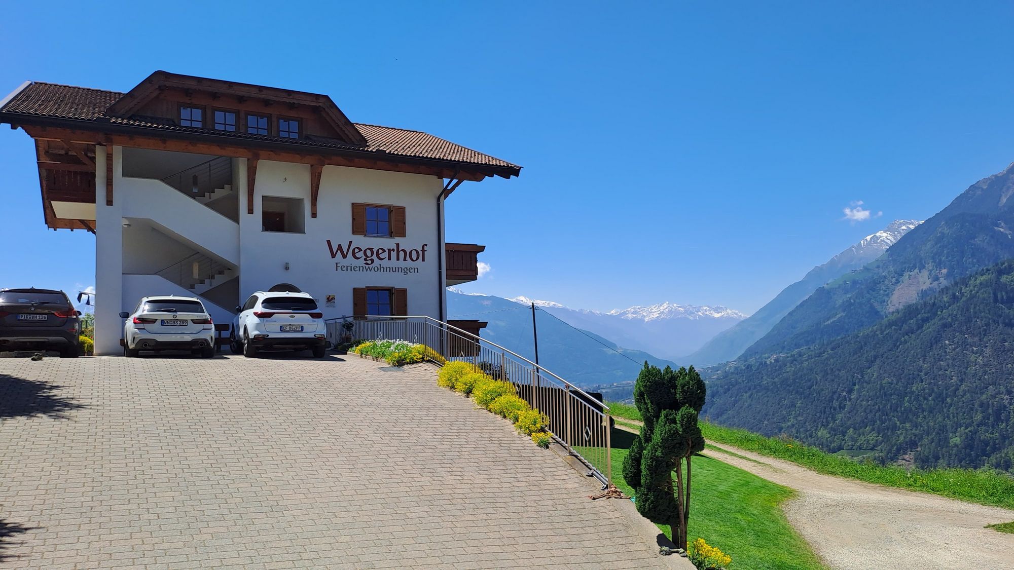 Ein weißes Ferienhaus namens Wegerhof mit geparkten Autos auf einer gepflasterten Auffahrt vor einem weiten Panorama aus Alpen mit schneebedeckten Gipfeln und strahlend blauem Himmel.